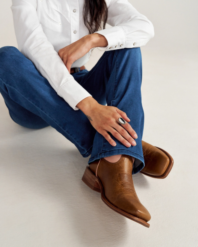 Person wearing a white shirt and blue jeans, seated on the floor, showing brown square-toed cowboy boots.