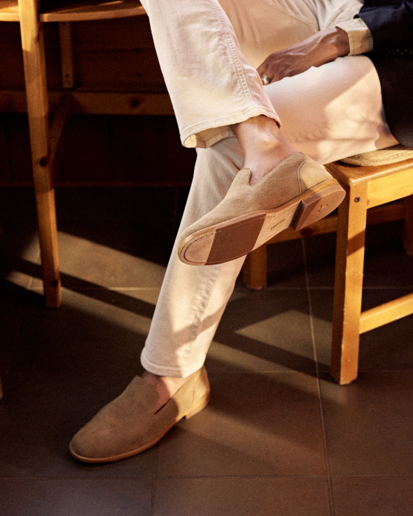 The Monterrey in Granite Suede. Person wearing light-colored trousers and Granite Suede loafers, sitting with one foot raised to show the sole.