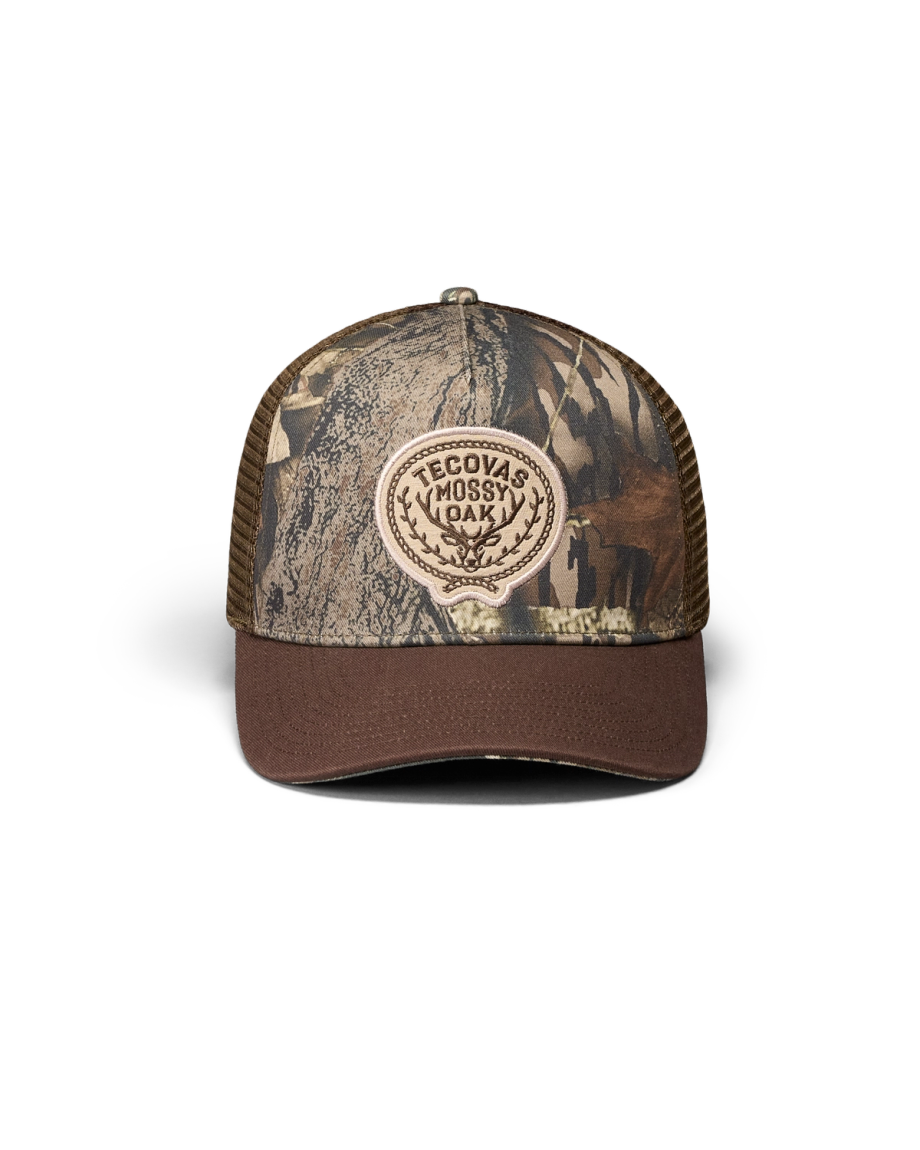 A camouflage-patterned trucker hat with a mesh back and a front patch that reads “Tecovas Mossy Oak.”.