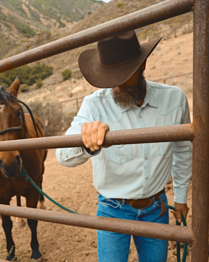 A man in a cowboy hat and denim clothes leans on a metal fence, holding a horse's reins, with a rural landscape in the background.