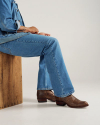 A person wearing blue jeans and brown leather boots sits on a wooden box against a plain background.