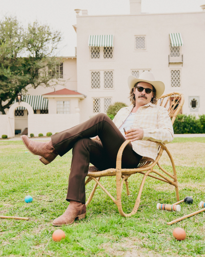 A person in a cowboy hat and sunglasses sits in a wicker chair on a lawn, holding a drink. Croquet equipment is scattered around, with a large white building in the background.