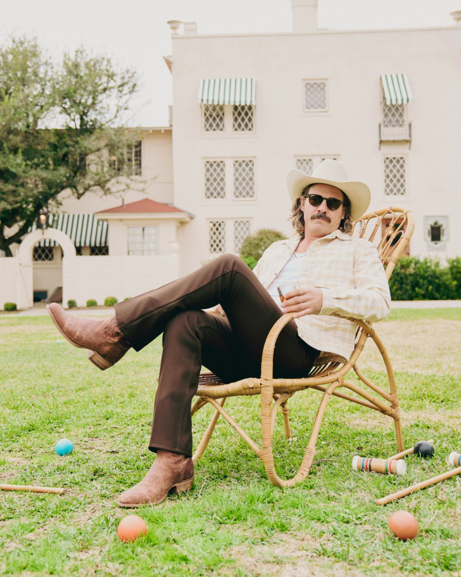 A man in a cowboy hat, boots, and sunglasses sits in a wicker chair on a lawn. Croquet equipment is scattered around.