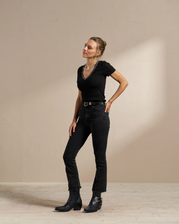 A woman stands against a plain background wearing a black short-sleeve top, black jeans, black belt, black boots, and a necklace. Her hair is tied back, and she is looking slightly to the side.