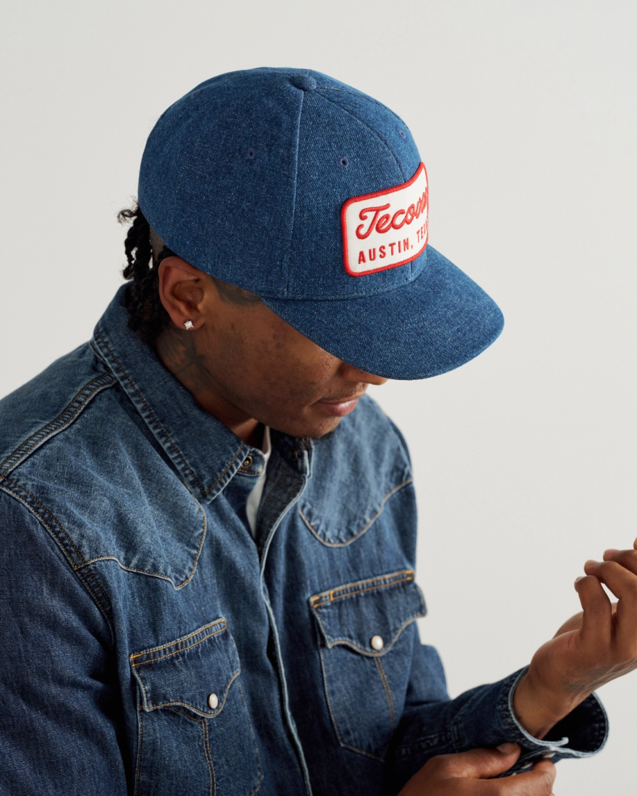 A person wearing a denim jacket and a blue cap with a red and white patch reading "Tecom Austin, TX" looks down while adjusting their sleeve.