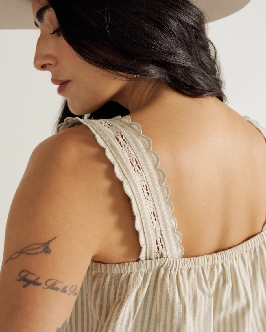 Woman with long dark hair wearing a sleeveless striped dress and a hat, showing a tattoo on her upper arm and decorative scalloped straps on her dress.