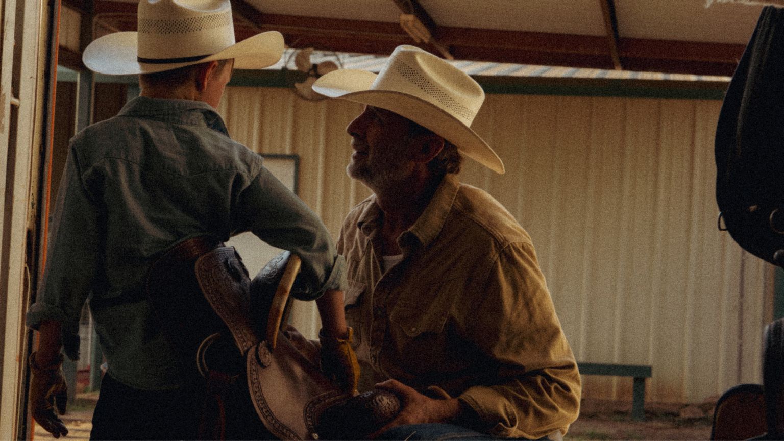 An adult and a child wearing cowboy hats talk indoors; the child holds a saddle while the adult looks up at him.