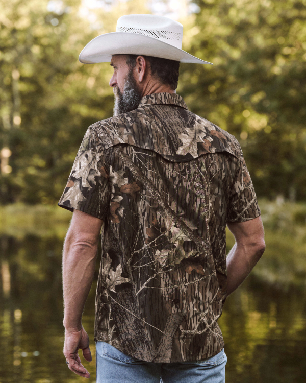 A man in a cowboy hat and short-sleeve camouflage shirt stands outdoors near water, facing away from the camera, with trees in the background.