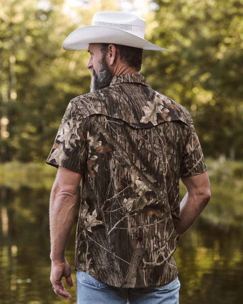 A man in a cowboy hat and short-sleeve camouflage shirt stands outdoors near water, facing away from the camera, with trees in the background.