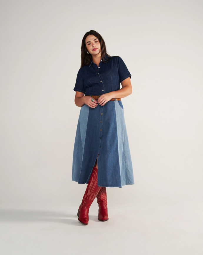 The Abby Wide Calf in Crimson Cowhide. Woman stands against a plain background wearing a short-sleeve denim shirt, two-tone button-front denim skirt, and striking crimson boots.