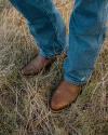 A person wearing blue jeans and brown leather cowboy boots stands on dry grass.