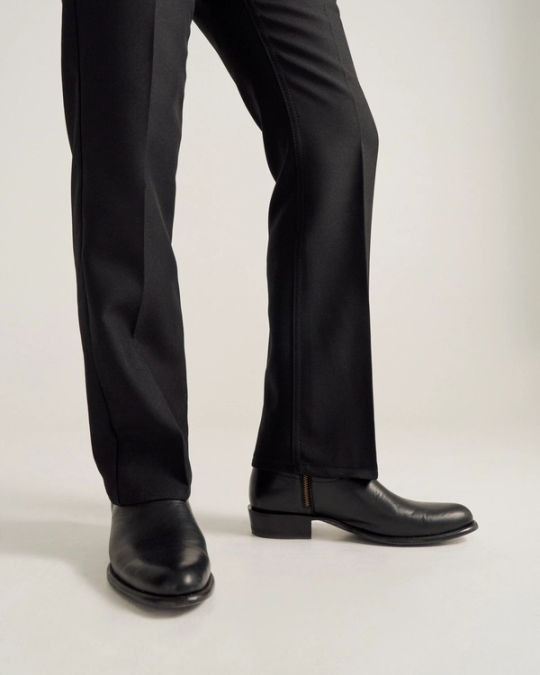A person wearing black dress pants and black leather shoes stands on a light-colored surface.