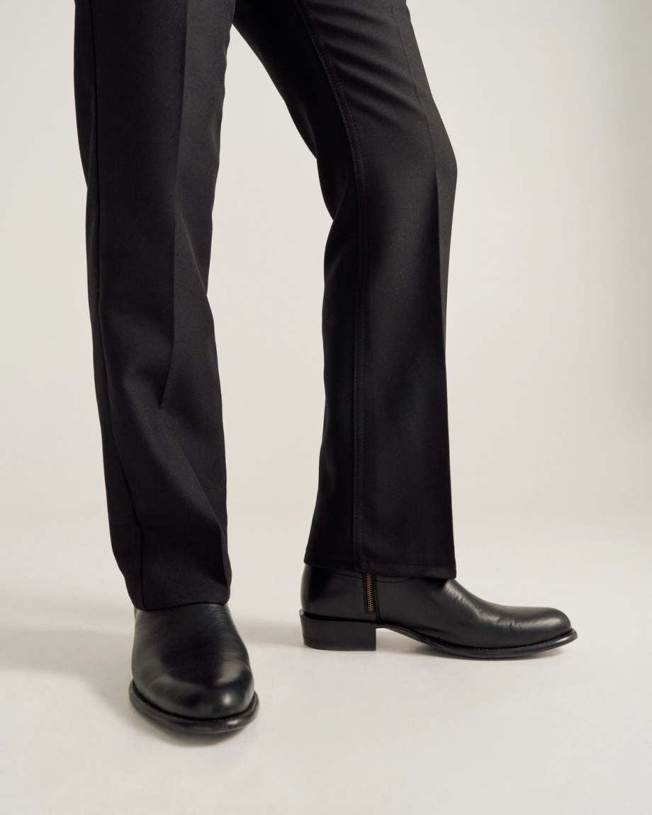 A person wearing black dress pants and black leather shoes stands on a light-colored surface.