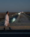 A man in a white robe and black boots walks along the side of a highway at dusk, with blurred car headlights in the background.