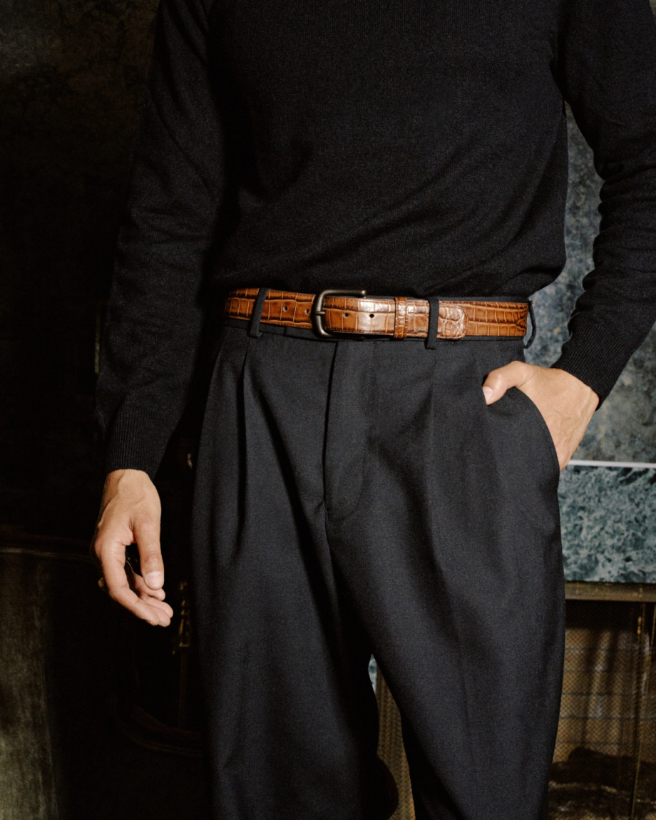 A person wearing a black sweater, pleated trousers, and a brown embossed leather belt.