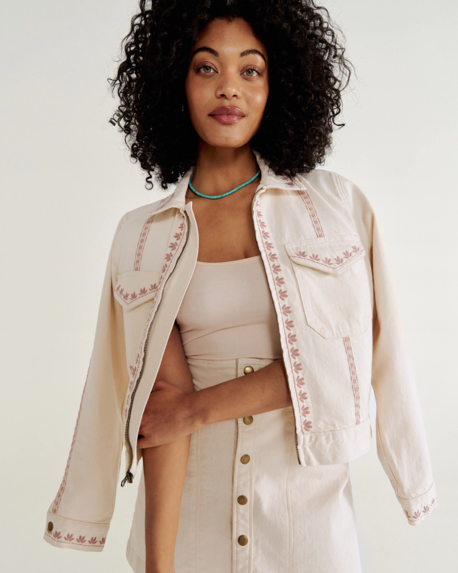 A woman with curly hair wears a light-colored jacket with embroidered patterns and a matching skirt. She has a turquoise necklace and stands against a plain background Natural Denim .