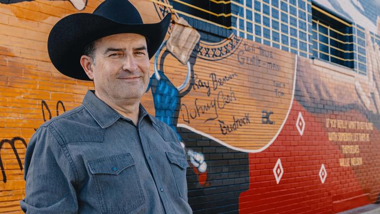 Image of a man wearing cowboy hat in front of a large mural. 