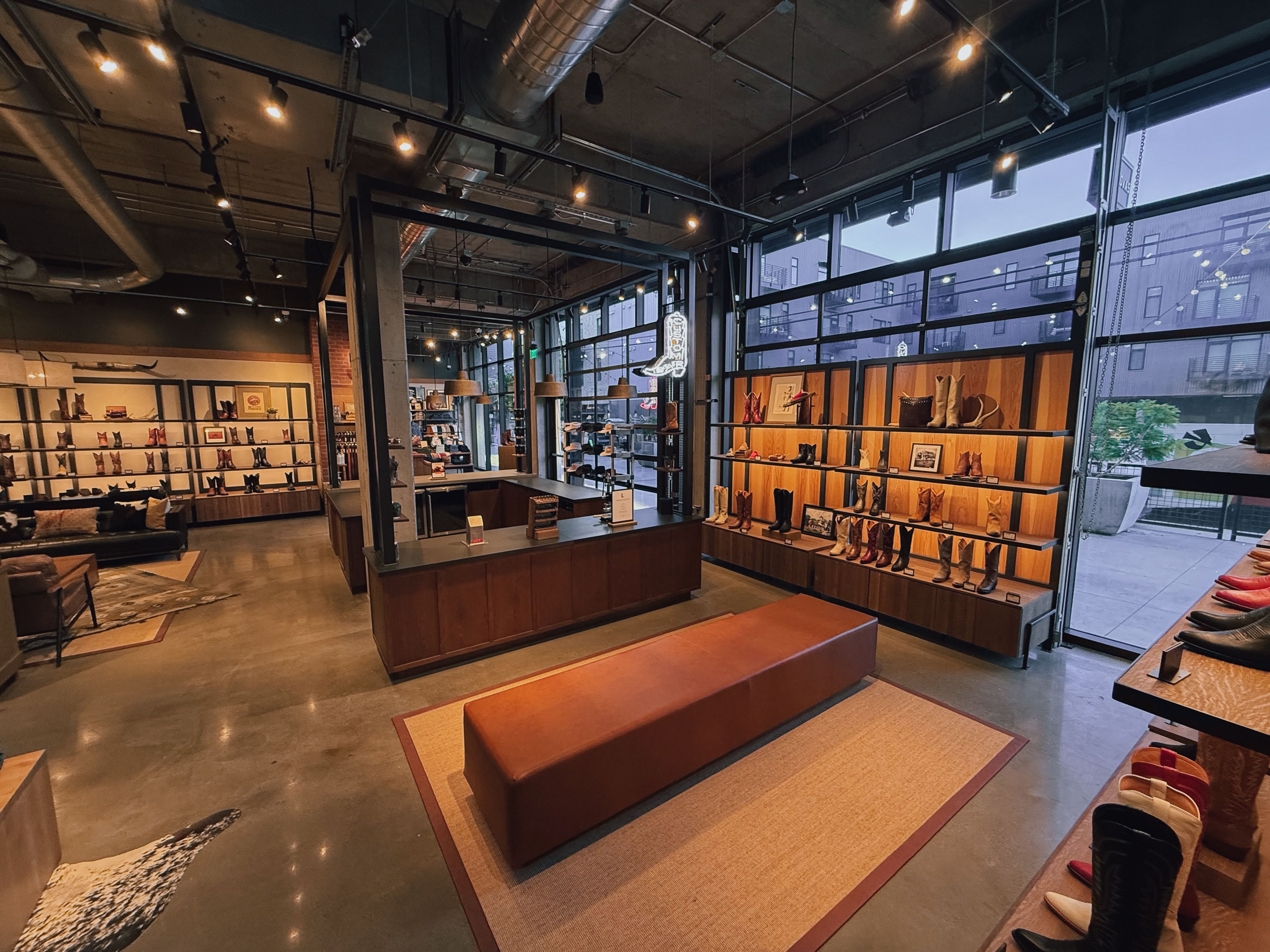 Tecovas store interior with leather seating, display shelves of boots and shoes, large windows, and industrial-style lighting and fixtures.