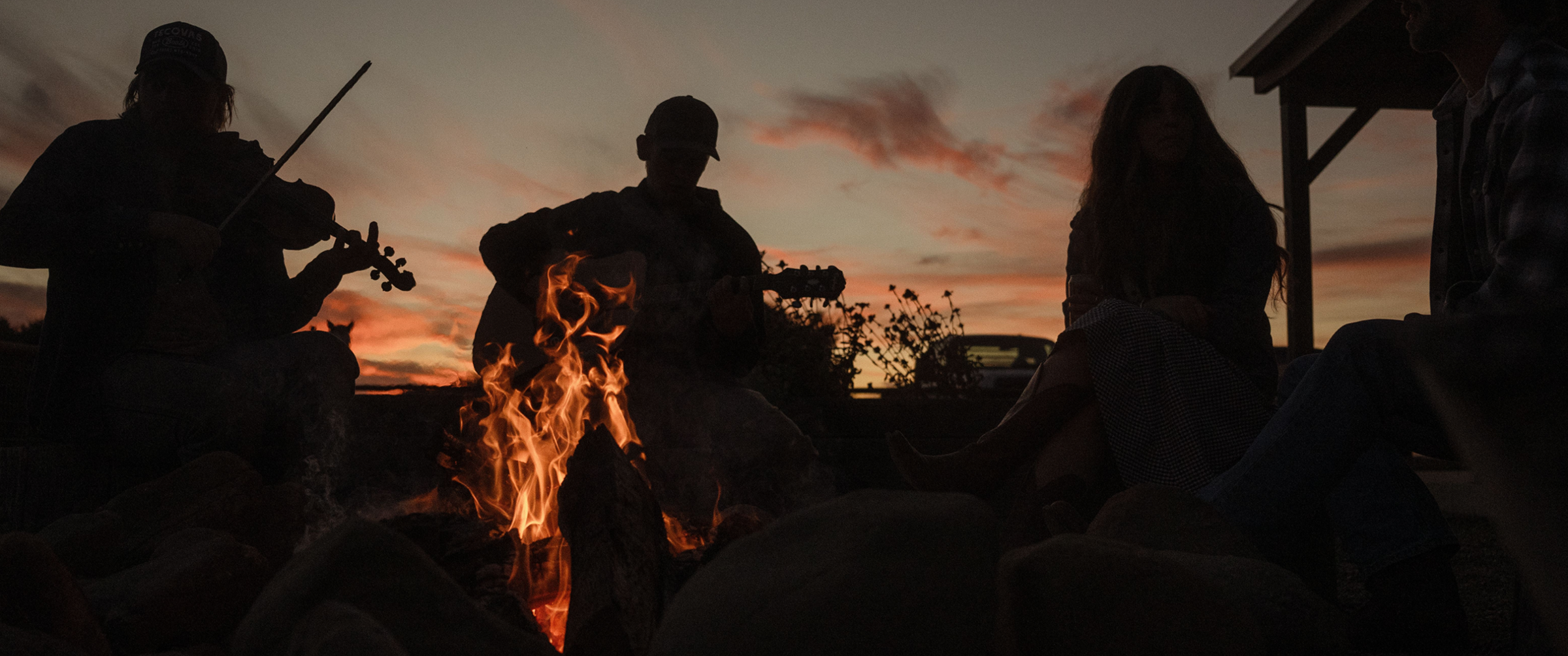 Silhouettes of people sitting around a campfire at sunset, with one person playing a violin and another playing a guitar.