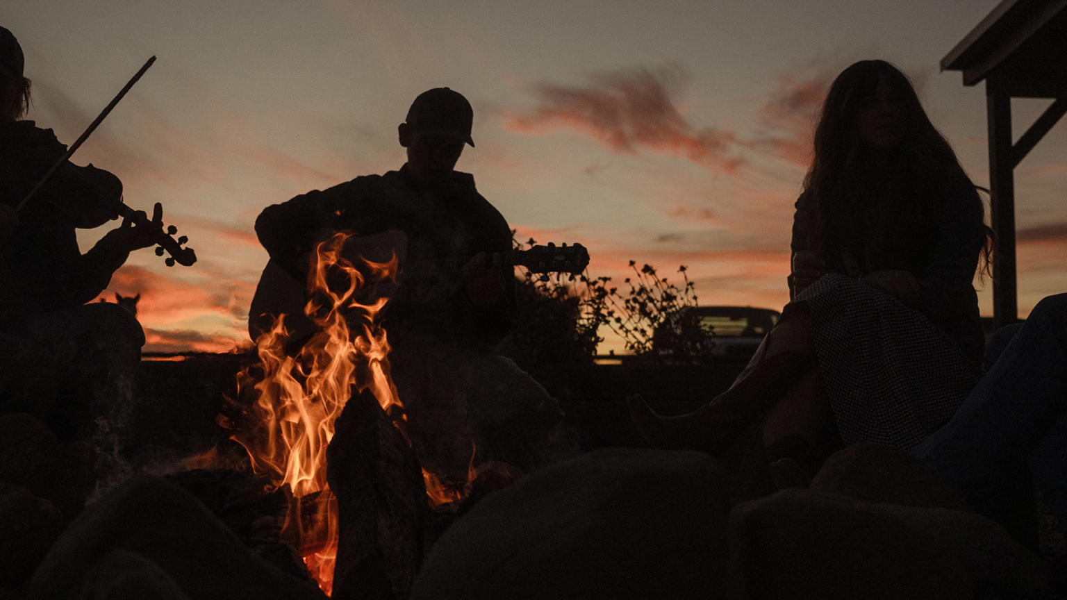 Silhouettes of people sitting around a campfire at sunset, with one person playing a violin and another playing a guitar.