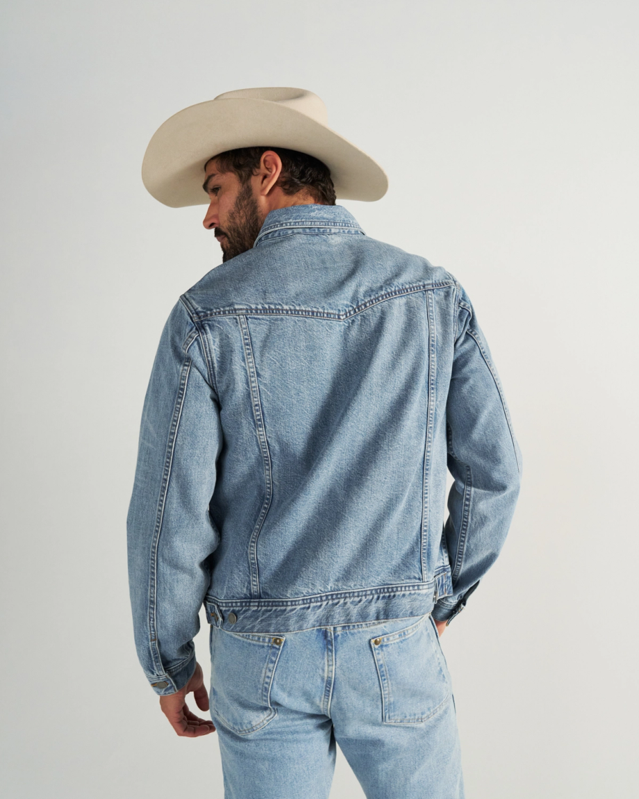 A man wearing a light denim jacket, matching jeans, and a beige cowboy hat stands facing away from the camera against a plain background.