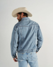 A man wearing a light denim jacket, matching jeans, and a beige cowboy hat stands facing away from the camera against a plain background.