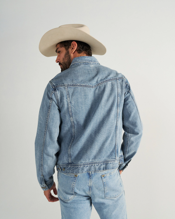 A man wearing a light denim jacket, matching jeans, and a beige cowboy hat stands facing away from the camera against a plain background.