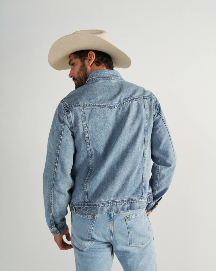 A man wearing a light denim jacket, matching jeans, and a beige cowboy hat stands facing away from the camera against a plain background.
