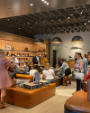A group of people shop and try on boots in a store with brown leather seating, wooden shelves, and boots displayed along the wall.