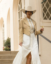 A woman in a white dress and beige fringe jacket stands on steps, holding a glass of champagne, wearing a beige hat. White arched windows are in the background.