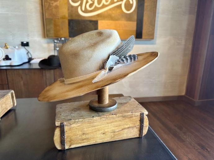 A rustic cowboy hat with a brown stain and braided band is displayed on a table, along with feathers and two more hats, in a store.