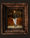 A man in a black shirt and light jeans stands in front of wooden shelves filled with boots, framed by an ornate, dark decorative frame.