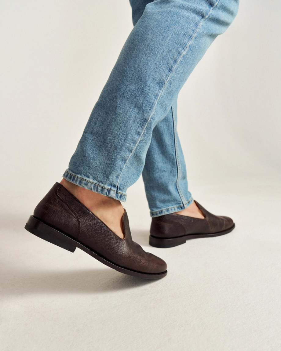 Person wearing light blue jeans and dark brown leather slip-on shoes walking on a light-colored surface.
