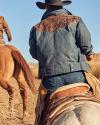 Two cowboys on horseback riding through a dry, grassy terrain, viewed from behind, with focus on the nearest rider's leather saddle.