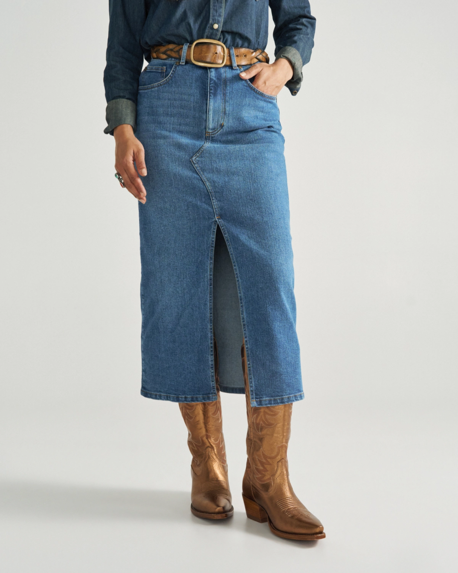 A person wearing a blue denim skirt with a front slit, a brown belt, a blue shirt, and brown cowboy boots stands against a plain background.