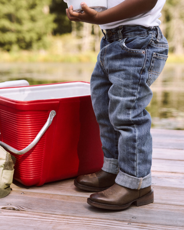 A child in jeans and boots stands next to a red cooler on a wooden dock by the water, holding a container.
