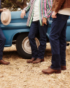 Three people in jeans and boots stand next to a blue truck. One holds a hat. Only their lower bodies are visible.