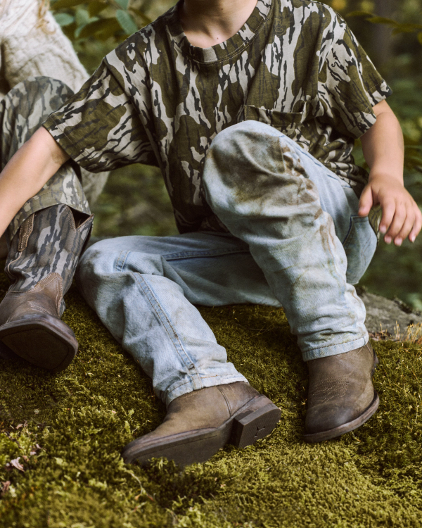 Two children sit on mossy ground outdoors, wearing camouflage shirts, light blue jeans, and brown boots. Only their bodies and partial faces are visible.