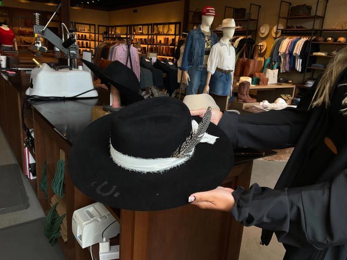 Person holding a black wide-brimmed hat with a white band and feather in a clothing store with hats, shirts, and mannequins in the background.