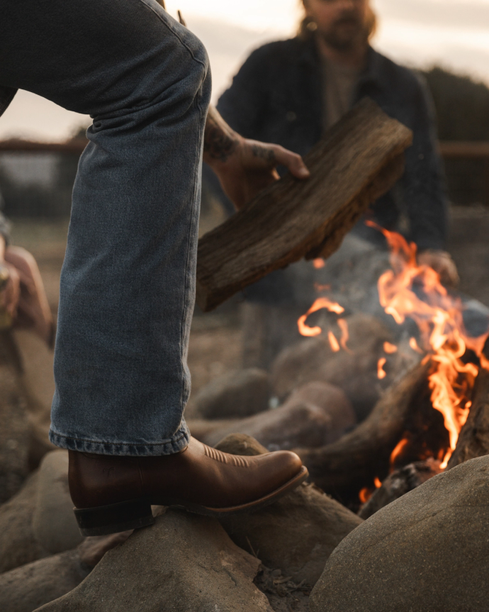 Person wearing jeans and brown boots adds a log to a campfire, with another person blurred in the background.