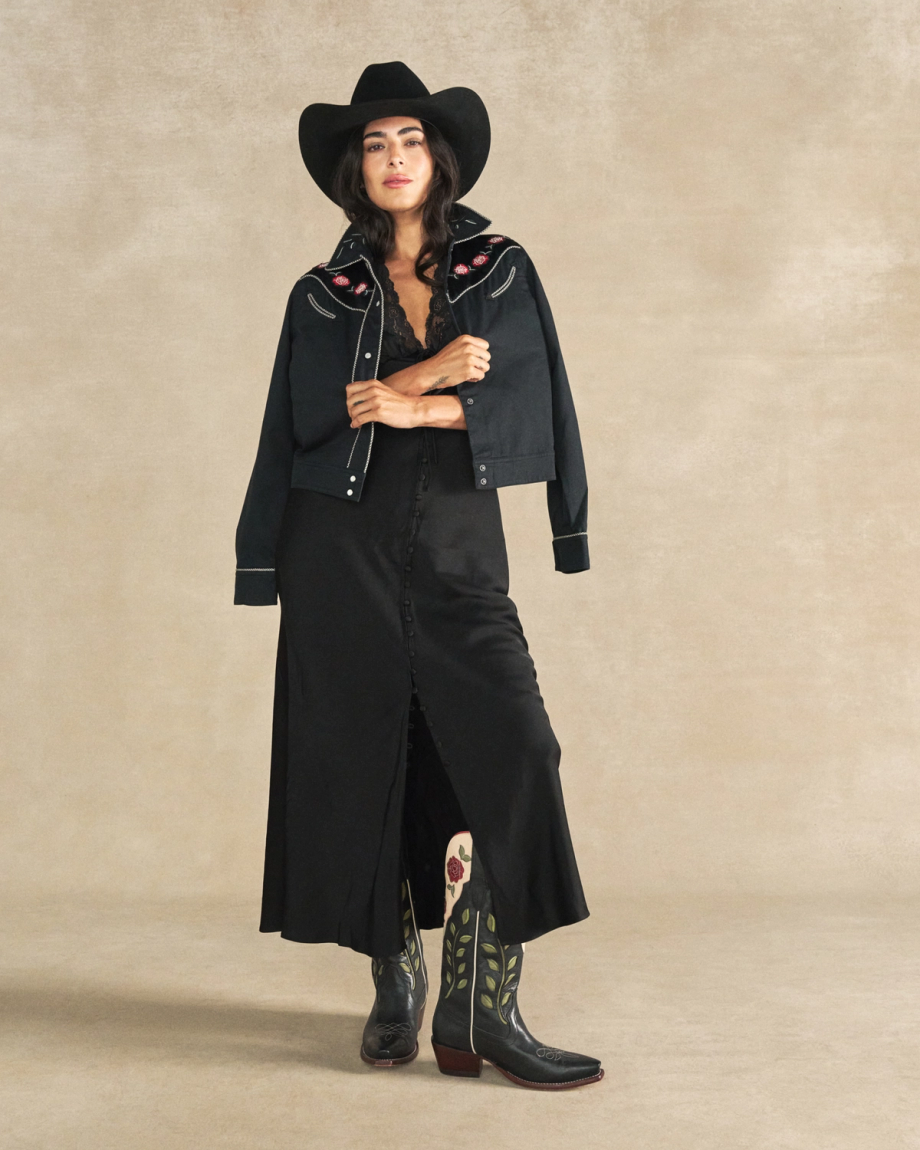 Woman in a black cowboy hat, embroidered jacket, long black skirt, and cowboy boots stands against a beige background, her look enhanced by The Annie Cowhide bag—a bold accent among midnight roses.