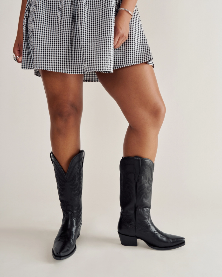 A person wearing a black and white checkered skirt and black cowboy boots stands against a plain background.