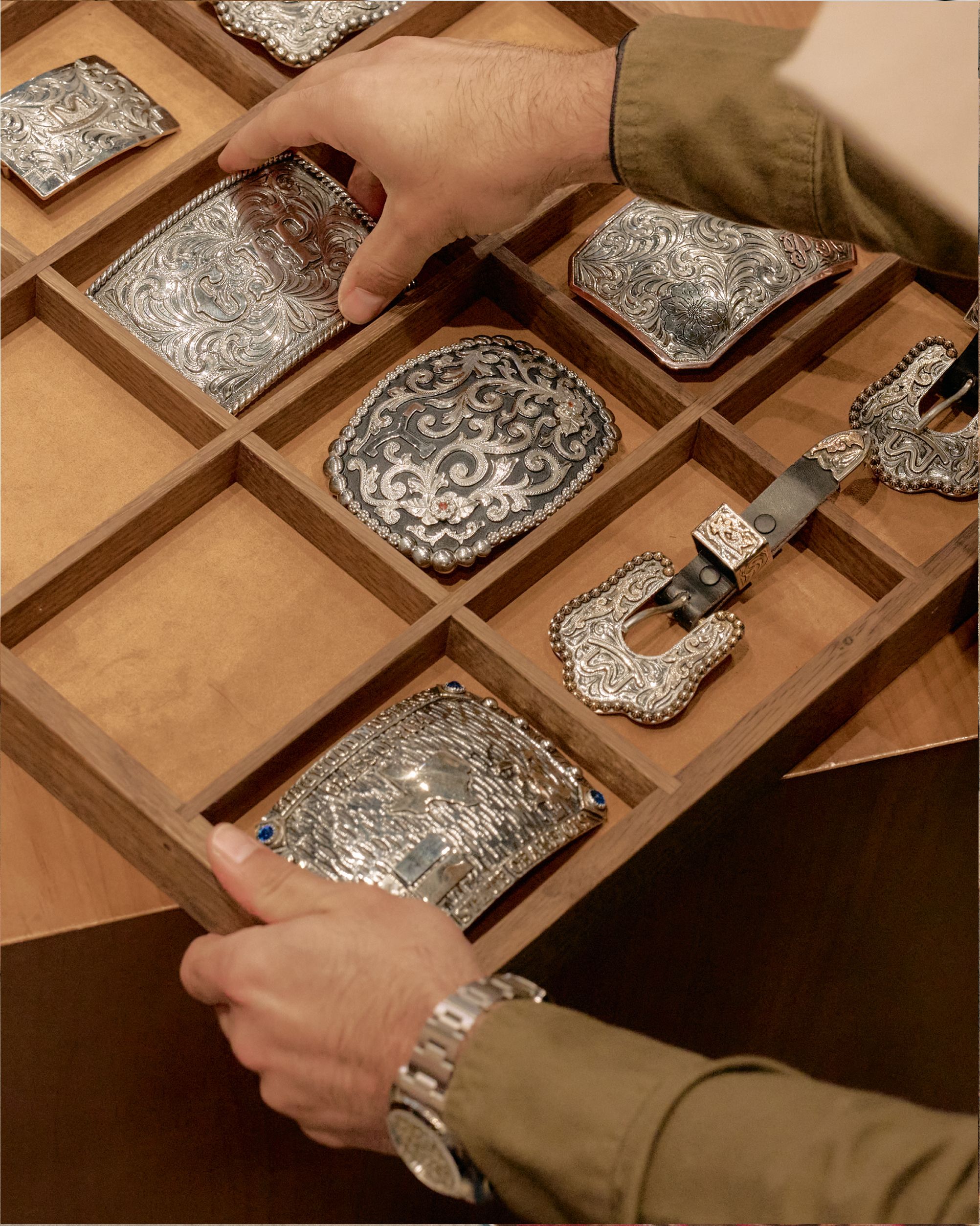 Person organizing a wooden drawer containing ornate silver belt buckles and accessories.