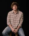 A young man with curly hair sits on a stool against a black background, wearing a plaid shirt, light jeans, and a wristwatch, looking directly at the camera.