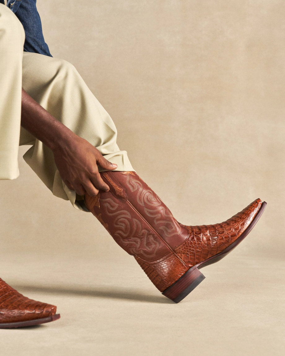 The Garrett in Pecan Hornback Caiman. Person wearing light pants pulls on a brown, patterned Pecan Hornback Caiman cowboy boot with a pointed toe.