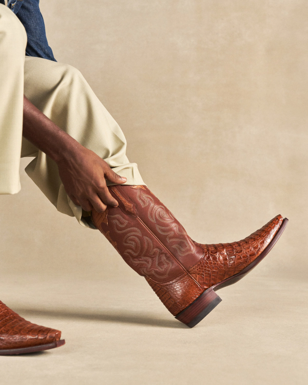 The Garrett in Pecan Hornback Caiman. Person wearing light pants pulls on a brown, patterned Pecan Hornback Caiman cowboy boot with a pointed toe.
