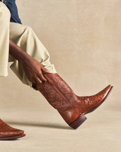 The Garrett in Pecan Hornback Caiman. Person wearing light pants pulls on a brown, patterned Pecan Hornback Caiman cowboy boot with a pointed toe.