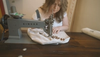 A person operates a sewing machine, stitching fabric with the word "HAND" partially visible. The sewing machine is labeled "SINGER." The person is focused on their work.