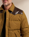 A mid-aged man wearing a mustard-colored padded jacket with brown leather patches on the shoulders and a green sweater underneath, partially visible against a plain background.