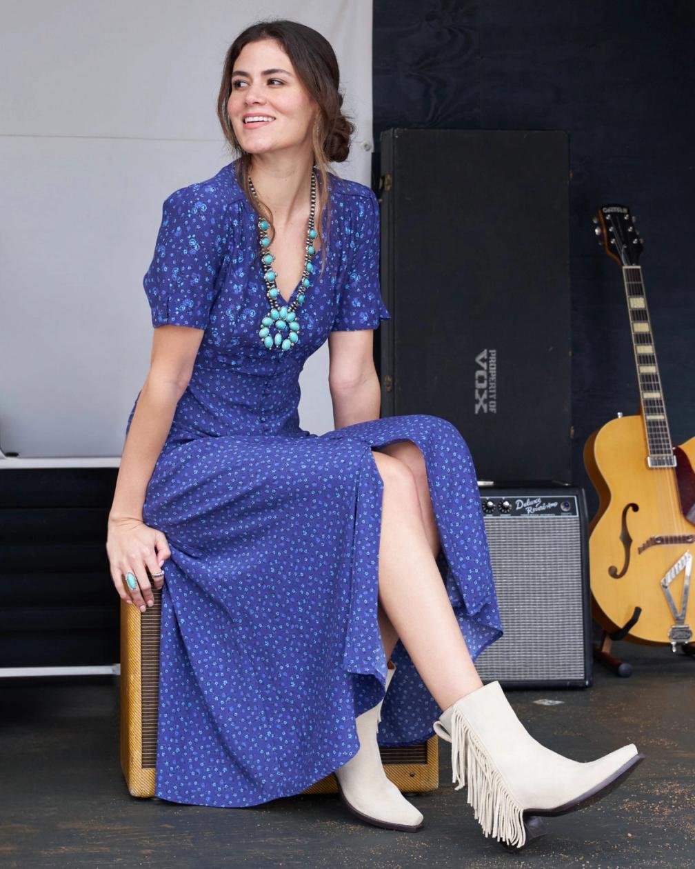 A woman in a blue dress and fringed boots sits on an amp, smiling, with a guitar and another amp in the background.